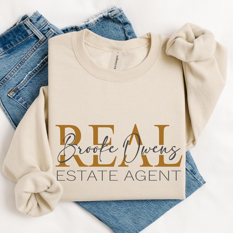 May include: A beige crewneck sweatshirt featuring the text REAL ESTATE AGENT in gold and black lettering, with the name Brooke Owens written in a black cursive script across the center. The sweatshirt is layered over a pair of blue denim jeans.