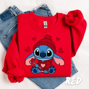 Peut inclure: Sweat-shirt rouge avec un dessin anim&eacute; de Stitch portant un chapeau rouge et tenant une sucette en forme de c&oelig;ur. Le sweat-shirt a un th&egrave;me de la Saint-Valentin avec des c&oelig;urs &eacute;parpill&eacute;s autour de Stitch.