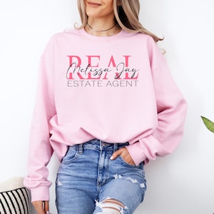 May include: A light pink crewneck sweatshirt featuring the text REAL in bold pink letters, the name Melissa Jay in a black cursive script, and the words ESTATE AGENT in black capital letters centered on the chest area of the garment.