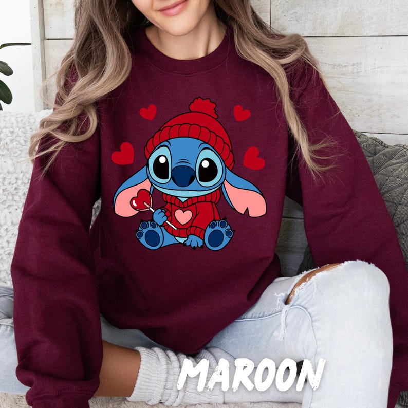 Peut inclure: Un sweat-shirt bordeaux avec un personnage de dessin anim&eacute; bleu tenant une sucette en forme de c&oelig;ur. Le personnage porte un chapeau et un pull rouge. Le sweat-shirt a un th&egrave;me de la Saint-Valentin avec des c&oelig;urs.