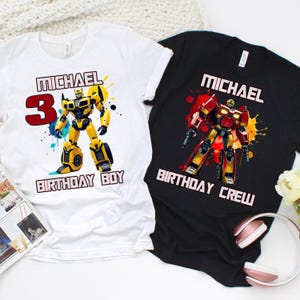 May include: Two t-shirts, one white and one black, featuring robot graphics. The white shirt has a yellow robot with the text "MICHAEL 3 BIRTHDAY BOY." The black shirt displays a red robot with the text "MICHAEL BIRTHDAY CREW."