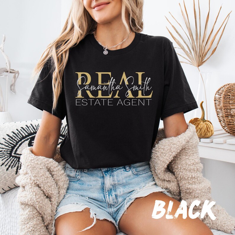May include: A black short-sleeved t-shirt featuring the gold text REAL, the white cursive script Samantha Smith, and the white text ESTATE AGENT printed on the front. The shirt has a crew neckline and is worn with light blue denim shorts.