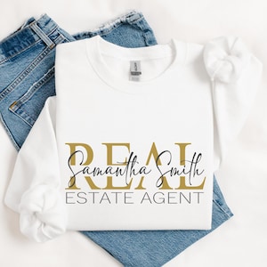 May include: A white long-sleeve sweatshirt featuring the gold text REAL, the black cursive name Samantha Smith, and the black text ESTATE AGENT printed on the front. The garment has a crew neckline and is paired with blue denim jeans.