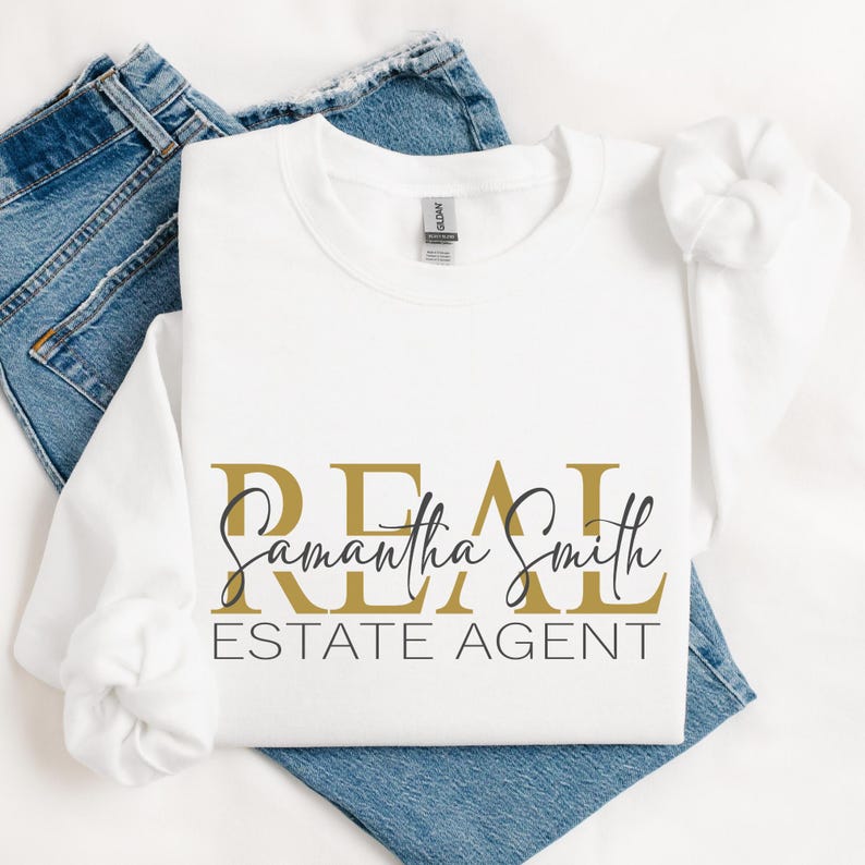 May include: A white long-sleeve sweatshirt featuring the gold text REAL, the black cursive name Samantha Smith, and the black text ESTATE AGENT printed on the front. The garment has a crew neckline and is paired with blue denim jeans.