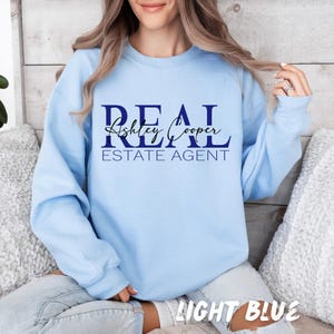 May include: A light blue crewneck sweatshirt featuring the dark blue text REAL ESTATE AGENT in a bold serif font, with the name Ashley Cooper written in a black cursive script layered across the center of the word REAL.