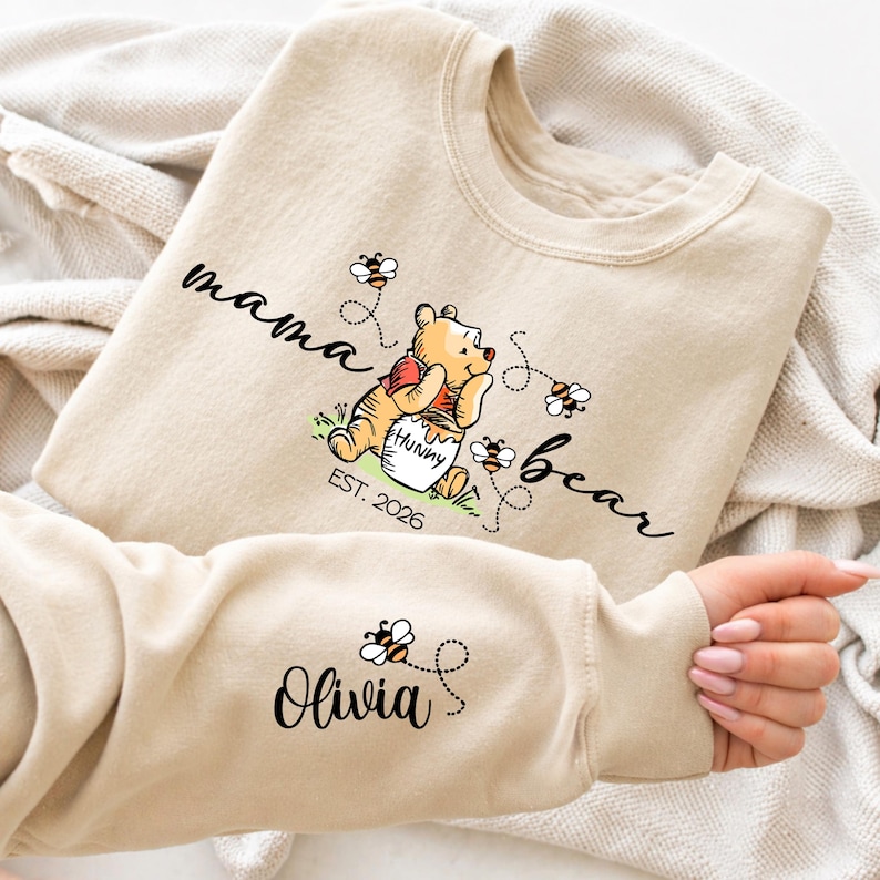 Peut inclure: Sweat-shirt beige avec les mots "mama bear" et "bear" en &eacute;criture cursive. Un ours de dessin anim&eacute; tient un pot de miel avec le mot "Hunny" dessus. La manche porte le nom "Olivia" et un motif d'abeille.