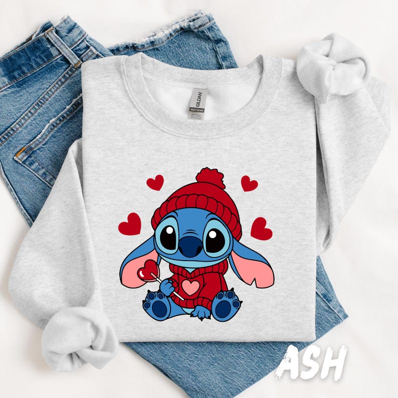 Peut inclure: Un sweat-shirt gris avec un personnage de dessin anim&eacute; bleu et rouge portant un chapeau rouge et tenant une sucette rouge. Le personnage est entour&eacute; de c&oelig;urs rouges. Le texte "ASH" est visible dans le coin inf&eacute;rieur droit de l'image.