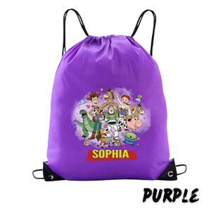May include: A purple drawstring backpack featuring a colorful graphic of Toy Story characters. The backpack has black drawstrings and a personalized name, "SOPHIA", on a red banner. The word "PURPLE" is printed in black at the bottom.
