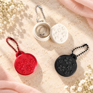 May include: Three circular, lidded containers with ball chain keychains in red, white, and black. Each container features a raised floral pattern on the lid. The white container is open, revealing a thin metal ring resting inside the base.