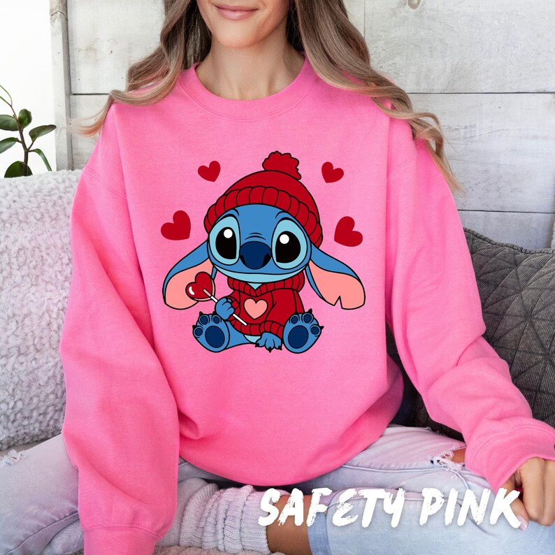 Peut inclure: Un sweat-shirt rose avec un personnage de dessin anim&eacute; bleu portant un chapeau rouge et tenant une sucette en forme de c&oelig;ur. Le personnage est entour&eacute; de c&oelig;urs rouges. Le texte "SAFETY PINK" est visible en bas de l'image.