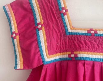 May include: A close-up of a bright pink dress with a square neckline and short sleeves. The neckline and sleeve edges are trimmed with colorful bands of white, blue, yellow, and pink. The dress is hanging on a white hanger.
