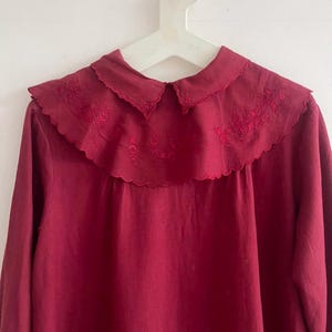 Puede incluir: Una blusa de color burdeos con cuello festoneado. El cuello presenta detalles florales bordados. La parte superior de manga larga está colgada de una percha blanca sobre un fondo blanco. La blusa está hecha de una tela suave y fluida.