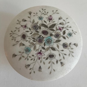 Puede incluir: Una tapa redonda de color crema con un diseño floral. El arreglo incluye racimos de flores en tonos azules, rosas y grises, con hojas y tallos verdes. La tapa tiene una superficie ligeramente texturizada.