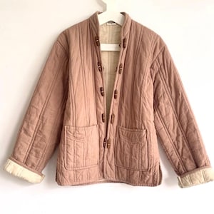 May include: A quilted jacket in a muted rose colour with a mandarin collar and wooden toggle closures. The jacket features two front pockets and cream-coloured cuffs. The jacket is hanging on a white hanger against a white background.