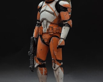 May include: A detailed action figure of a clone trooper in orange and white armor, holding a black blaster rifle. The figure has a helmet, chest armor, and leg armor. The background is black.