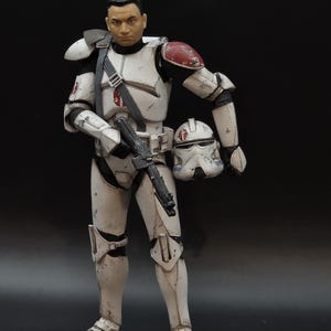 May include: A detailed action figure of a character in white and black armor with red accents, holding a black blaster rifle and a helmet. The figure has a realistic face and is posed against a dark background. The armor shows signs of wear.
