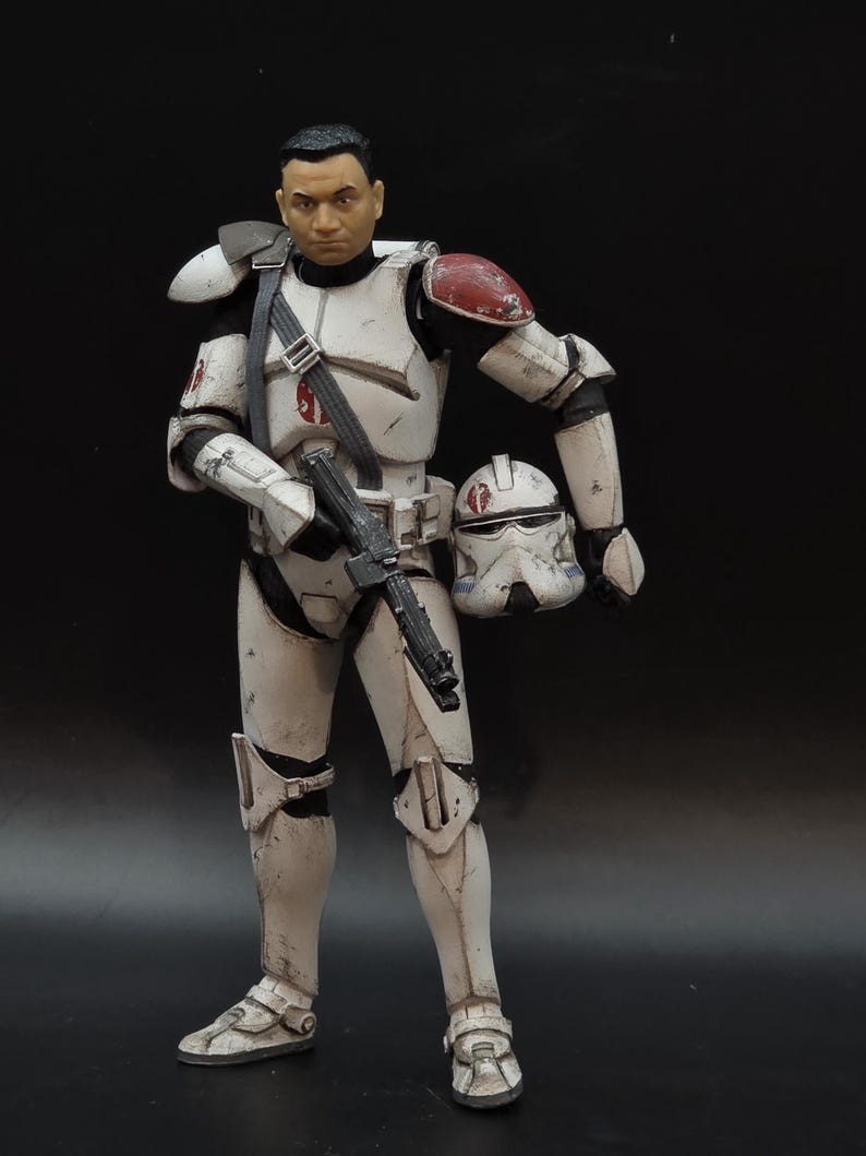 May include: A detailed action figure of a character in white and black armor with red accents, holding a black blaster rifle and a helmet. The figure has a realistic face and is posed against a dark background. The armor shows signs of wear.