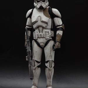以下が含まれることがあります： スターウォーズシリーズのキャラクターであるストームトルーパーの、風化した白とグレーのアクションフィギュア。ブラスターライフルを持ち、ヘルメットとアーマーには茶色と黒のアクセントがあり、暗い背景に立っています。