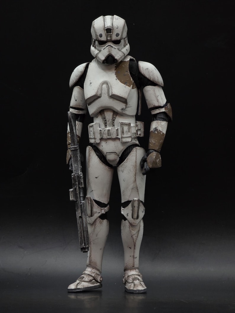 以下が含まれることがあります： スターウォーズシリーズのキャラクターであるストームトルーパーの、風化した白とグレーのアクションフィギュア。ブラスターライフルを持ち、ヘルメットとアーマーには茶色と黒のアクセントがあり、暗い背景に立っています。