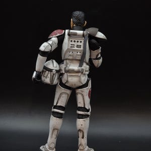 May include: A detailed action figure of a character in white and black armor, with red accents on the shoulders and a helmet in hand. The figure has a weathered appearance, suggesting battle damage. The background is a solid black.
