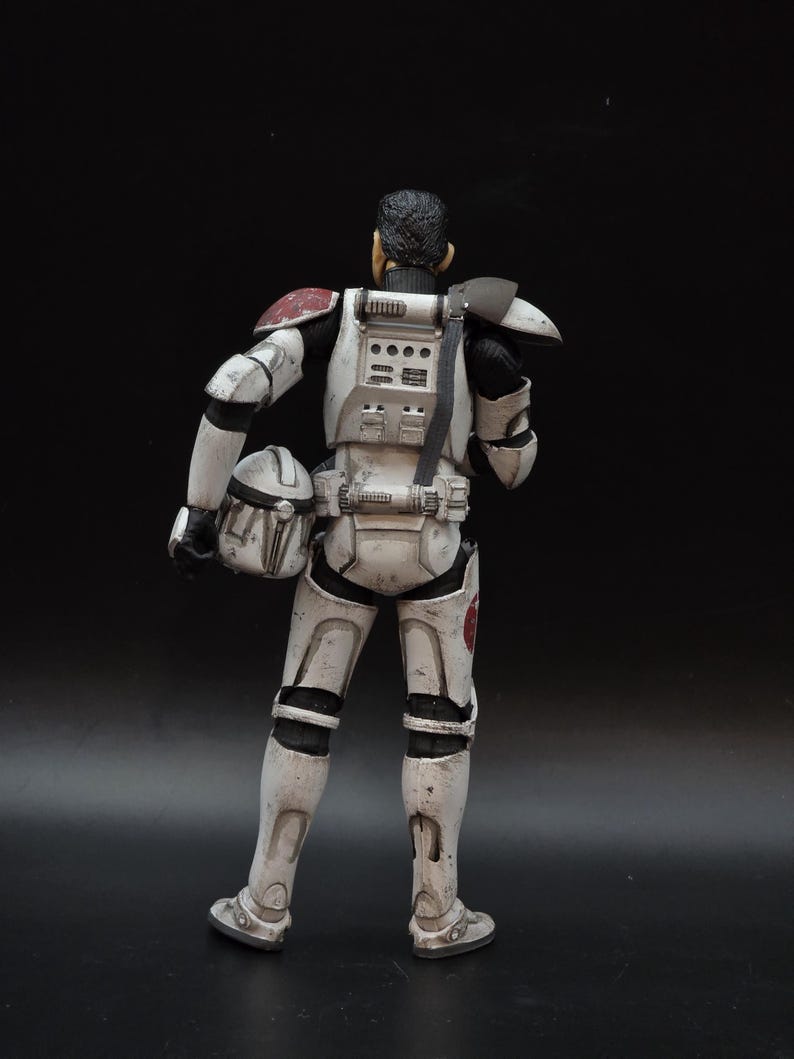 May include: A detailed action figure of a character in white and black armor, with red accents on the shoulders and a helmet in hand. The figure has a weathered appearance, suggesting battle damage. The background is a solid black.