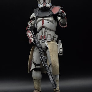May include: A detailed action figure of a clone trooper from Star Wars. The figure is predominantly white with red and black accents, holding a black rifle. It features a helmet, armor, and a tan-colored skirt. The figure is standing against a black background.