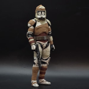 May include: A detailed action figure of a Star Wars clone trooper in a standing pose. The figure is predominantly white, with brown and tan accents on the armor and helmet. It is holding a black blaster rifle and has a brown belt.