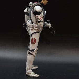 May include: A side profile of a white and gray action figure in a combat suit. The figure has a black head and a red emblem on the shoulder and leg. The figure is holding a black weapon.