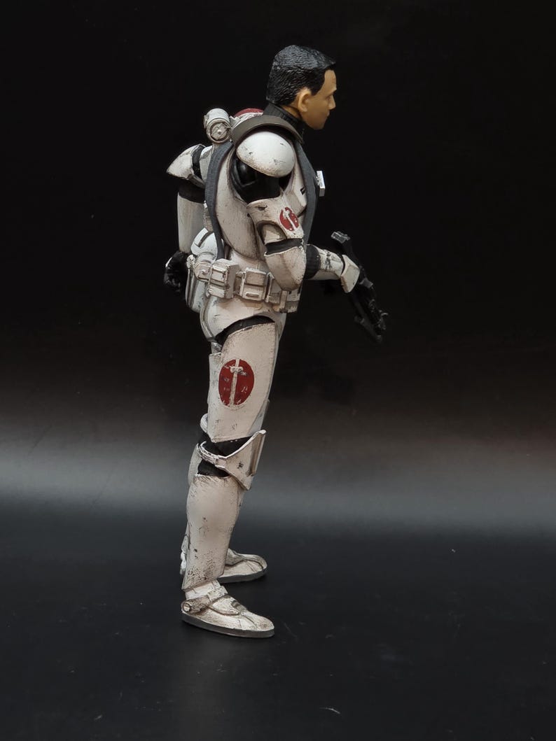 May include: A side profile of a white and gray action figure in a combat suit. The figure has a black head and a red emblem on the shoulder and leg. The figure is holding a black weapon.