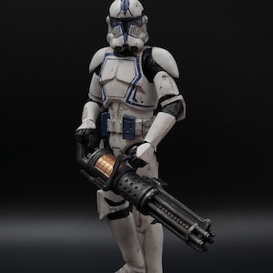 Könnte beinhalten: Eine weiß-blaue Actionfigur eines Soldaten in Rüstung, der eine große schwarze Waffe hält. Die Figur hat einen Helm und steht vor einem schwarzen Hintergrund. Die Rüstung hat blaue Akzente und die Waffe hat einen zylindrischen Lauf.