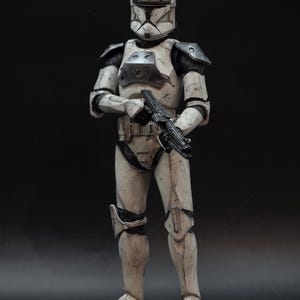 May include: A detailed action figure of a clone trooper in weathered white and gray armor, holding a black blaster rifle. The helmet features a green and white design. The figure stands against a dark background.