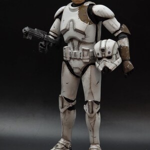 以下が含まれることがあります： 白いと茶色の鎧を着た兵士のアクションフィギュア。ブラスター銃とヘルメットを持っています。フィギュアはリアルな顔をしており、暗い背景にポーズをとっています。鎧には使用感が見られます。
