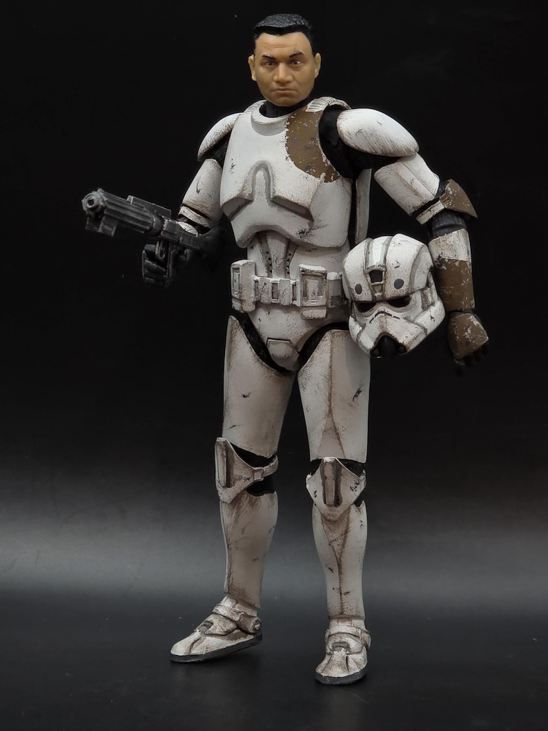 以下が含まれることがあります： 白いと茶色の鎧を着た兵士のアクションフィギュア。ブラスター銃とヘルメットを持っています。フィギュアはリアルな顔をしており、暗い背景にポーズをとっています。鎧には使用感が見られます。