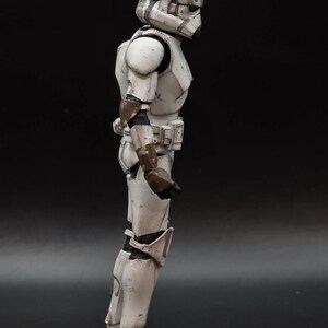 以下が含まれることがあります： 白とグレーのストームトルーパーのアクションフィギュアの側面図。フィギュアはヘルメット、アーマー、ブーツを着用しています。アーマーには風化や戦闘による損傷が見られます。フィギュアは黒い背景に立っています。