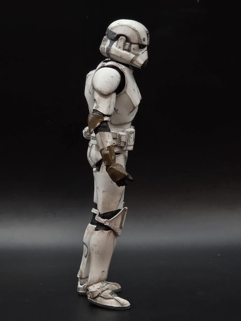 以下が含まれることがあります： 白とグレーのストームトルーパーのアクションフィギュアの側面図。フィギュアはヘルメット、アーマー、ブーツを着用しています。アーマーには風化や戦闘による損傷が見られます。フィギュアは黒い背景に立っています。