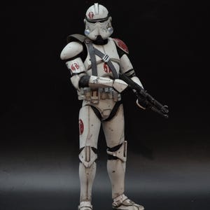 May include: A detailed action figure of a white and off-white armored soldier with red accents on the shoulder and helmet. The figure is holding a black rifle and has a utility belt. The figure is standing against a black background.