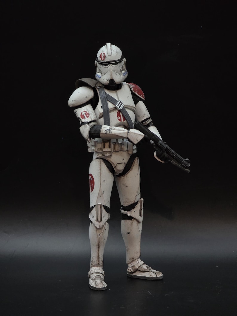 May include: A detailed action figure of a white and off-white armored soldier with red accents on the shoulder and helmet. The figure is holding a black rifle and has a utility belt. The figure is standing against a black background.
