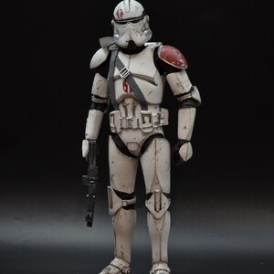 May include: A detailed action figure of a white and gray armored soldier with red shoulder accents. The figure is holding a black blaster rifle and has a helmet with a red marking. The figure is standing against a black background.
