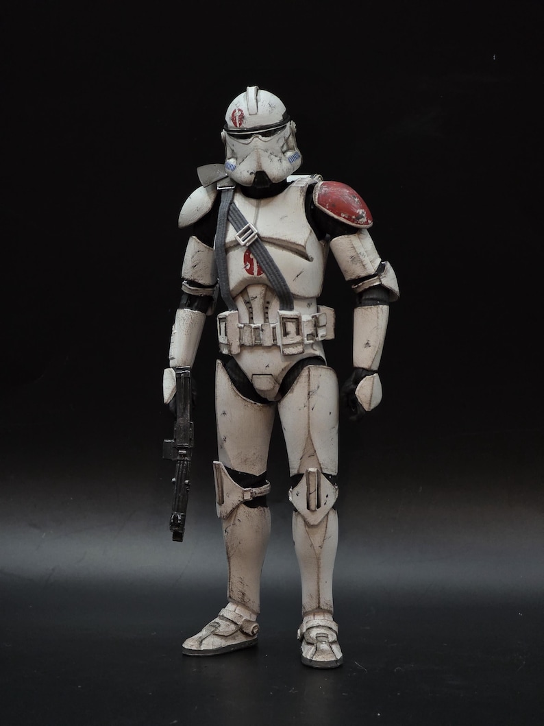 May include: A detailed action figure of a white and gray armored soldier with red shoulder accents. The figure is holding a black blaster rifle and has a helmet with a red marking. The figure is standing against a black background.