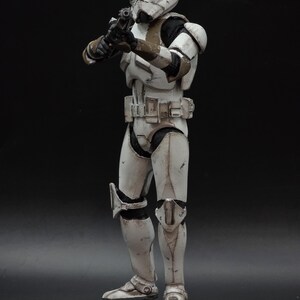 以下が含まれることがあります： スターウォーズのインペリアルストームトルーパーのアクションフィギュア。ブラスターを構え、戦闘態勢をとっている。白を基調とし、黒と茶色のアクセントが施され、暗い背景に配置されている。