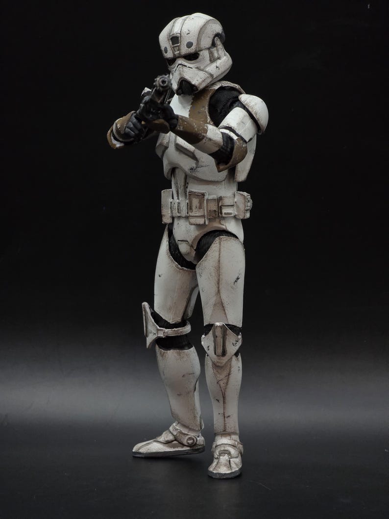以下が含まれることがあります： スターウォーズのインペリアルストームトルーパーのアクションフィギュア。ブラスターを構え、戦闘態勢をとっている。白を基調とし、黒と茶色のアクセントが施され、暗い背景に配置されている。