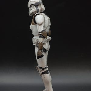以下が含まれることがあります： ストームトルーパー風のアーマーをまとった白とグレーのアクションフィギュアの側面図。フィギュアはヘルメット、肩パッド、脚部アーマーを備えています。アーマーは、茶色のアクセントで風化したディテールが施されています。フィギュアは暗い面に立っています。