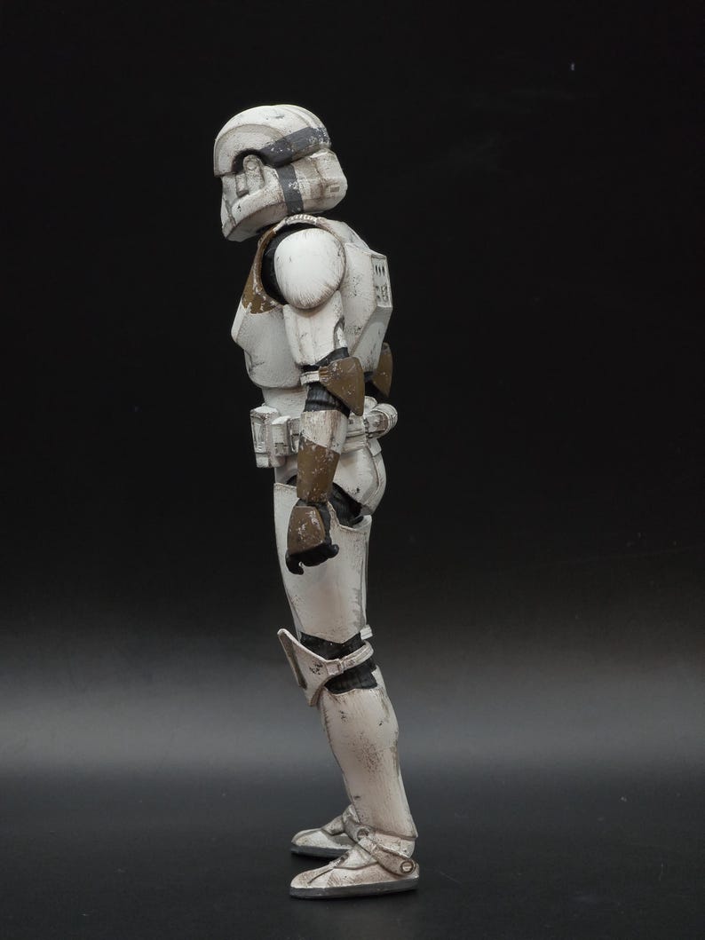 以下が含まれることがあります： ストームトルーパー風のアーマーをまとった白とグレーのアクションフィギュアの側面図。フィギュアはヘルメット、肩パッド、脚部アーマーを備えています。アーマーは、茶色のアクセントで風化したディテールが施されています。フィギュアは暗い面に立っています。
