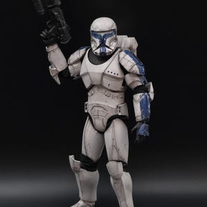 Pode incluir: Figura de ação branca e azul de um soldado em armadura, segurando uma pistola blaster preta. A figura tem um capacete branco com detalhes azuis e uma mochila. A armadura é detalhada com efeitos de desgaste.