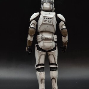 以下が含まれることがあります： 後ろから見た、白とグレーのアーマーを着た兵士のアクションフィギュア。フィギュアはヘルメット、肩パッド、ユーティリティベルトを備えています。アーマーには茶色のアクセントがあり、黒い手袋とブーツが付いています。フィギュアは暗い表面に立っています。