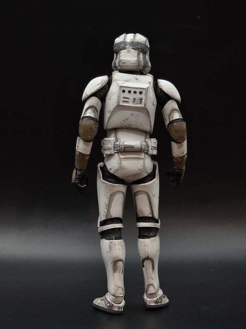 以下が含まれることがあります： 後ろから見た、白とグレーのアーマーを着た兵士のアクションフィギュア。フィギュアはヘルメット、肩パッド、ユーティリティベルトを備えています。アーマーには茶色のアクセントがあり、黒い手袋とブーツが付いています。フィギュアは暗い表面に立っています。