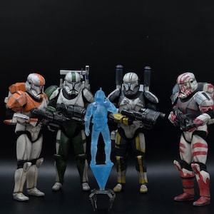 May include: Four action figures of armored soldiers with distinct colour schemes, including white, orange, green, and red. A blue holographic figure is in front. The figures are holding black rifles.