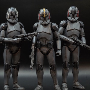 May include: Three grey action figures of soldiers in armour, each holding a black rifle. The figures have detailed helmets with red and yellow accents. The figures are standing in a row against a dark background.
