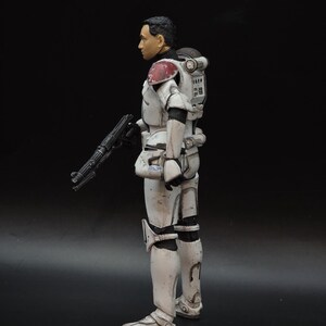May include: A white and gray action figure of a soldier in armor, holding a black rifle. The figure has a black helmet and a red and white shoulder pad. The figure is standing on a black surface.