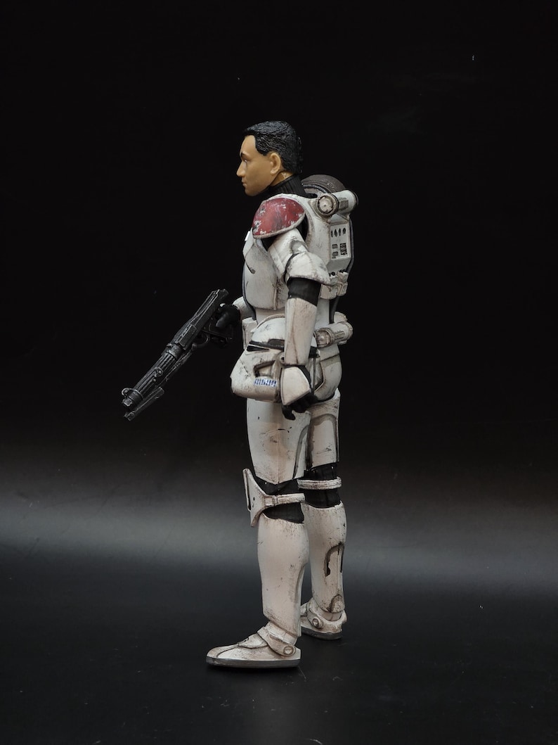 May include: A white and gray action figure of a soldier in armor, holding a black rifle. The figure has a black helmet and a red and white shoulder pad. The figure is standing on a black surface.